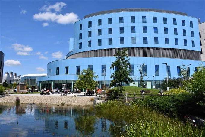 Image of blue building next to a pond with green grasses and trees. A blue sky with a few white clouds.