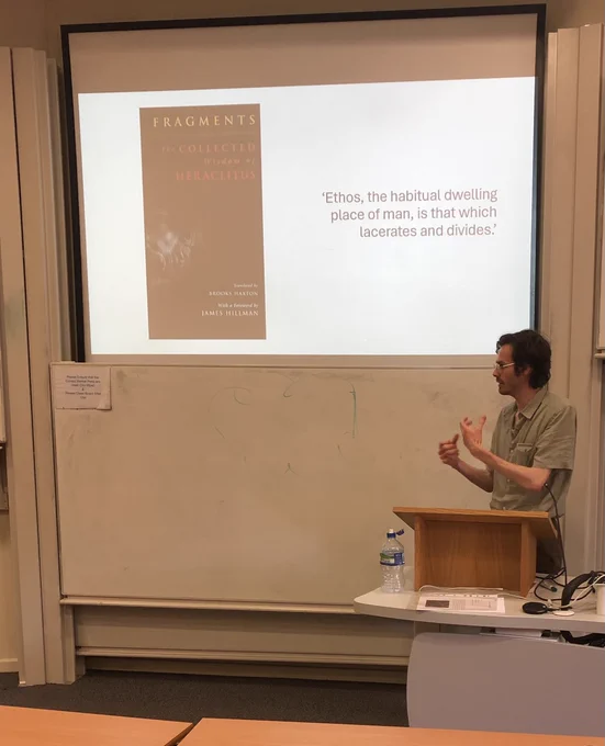 Photo of Jamie Davies speaking in front of a screen containing a quote from Heraclitus -- 'Ethos, the habitual dwelling place of man, is that which lacerates and divides'.