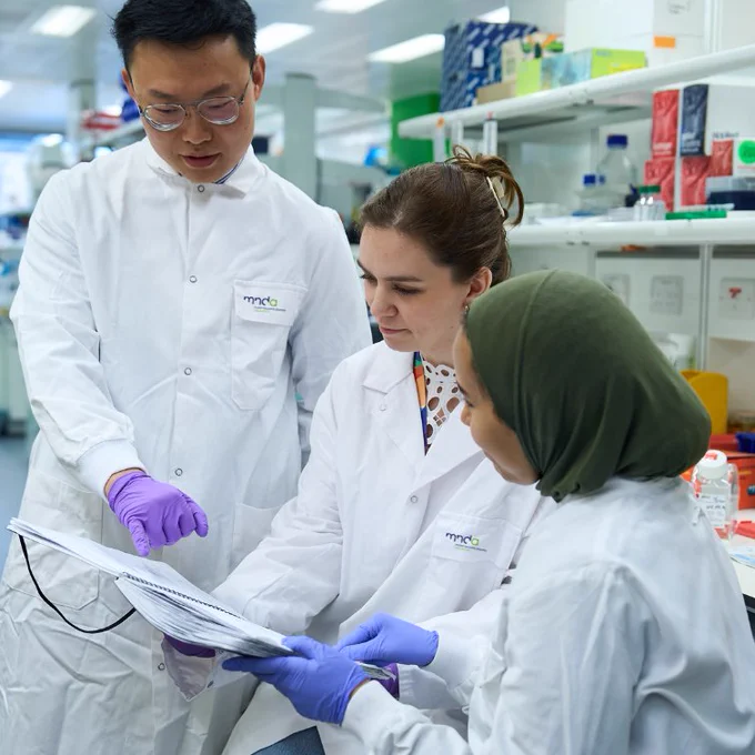 3 researchers wearing MND Association branded lab coats looking a book while in a laboratory