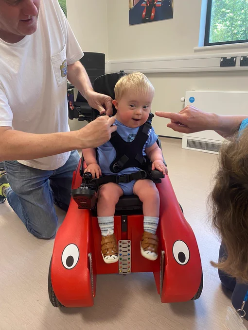 Morris smiling at the camera while people from Designability fit his harness round him in his Wizzybug powered wheelchair