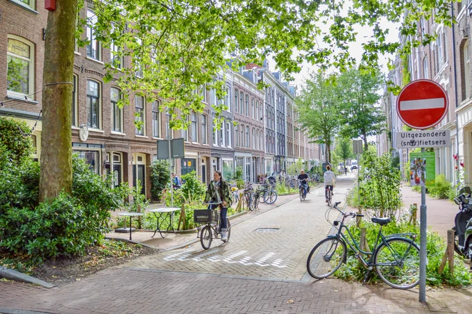 People walk and cycle through Amsterdam’s De Pijp neighborhood. Rather than cars in the on-street parking spaces, they are filled with trees, terraces, benches, playgrounds, and parked bikes.