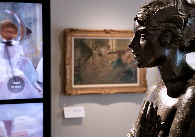 Photograph showing 3 objects in a gallery. On the left hand side is a tall digital screen, the image is obscured. Next to it on a grey wall is a painting in the impressionist style by Edgar Degas, The Rehearsal. The painting is not fully in focus and the details are hard to decipher. In the foreground, a black material sculpture is carved to resemble a woman with short curly hair, her hands are raised to her chest.