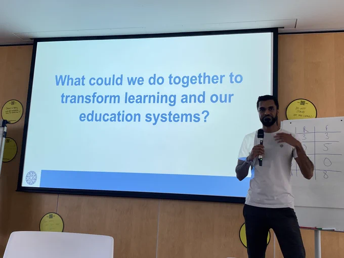 Male on white T-shirt in front of a slide that says “what could we do together to transform learning and our education systems’