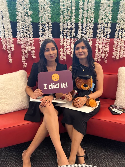 Two people sitting, smiling at the camera wearing crowns and their diplomas alongside a grad bear, holding a sign that says "I did it!"