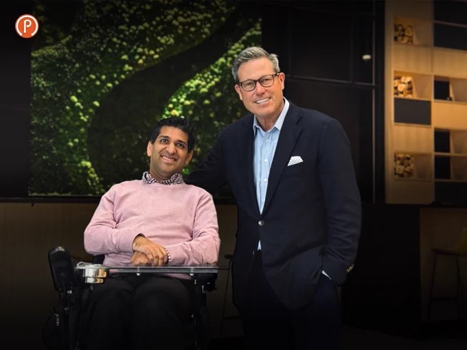 This is a picture of Nabeel Ramji, CEO of Pedesting with Scott Hutcheson, Executive chair of Aspen properties on the announcement of Ampersand and Edison buildings to the Pedesting app. Nabeel is in a wheelchair wearing a pink sweater and Scott standing wearing a blue suit.