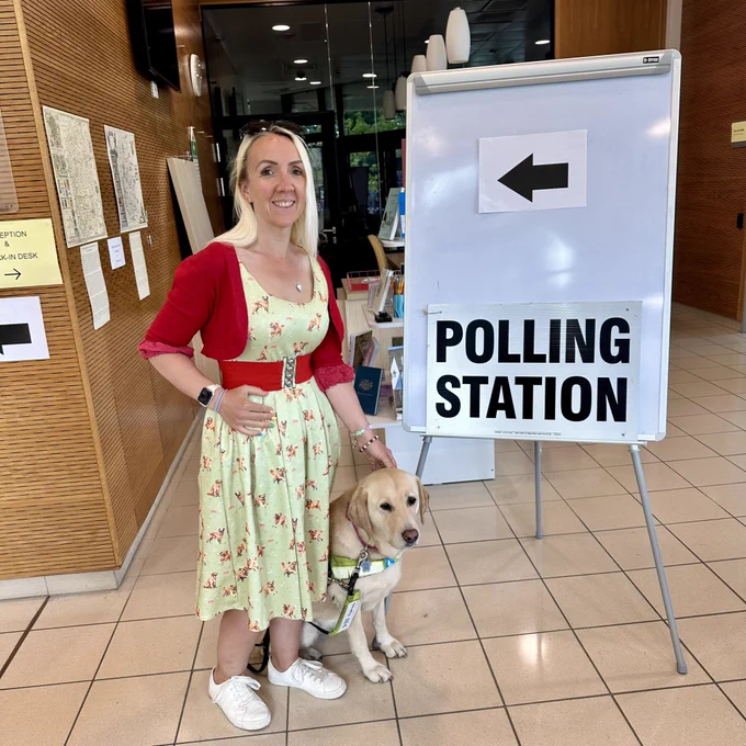 Photo shows me, a woman with blonde hair wearing a green dress and red cardigan, stood next to guide dog Ziggy, a yellow Labrador retriever. We are standing next to a sign that says ‘Polling Station’