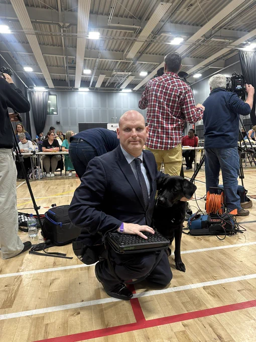 The picture shows me, Sean the human in a black suit and light blue shirt sitting next to Shawn the Shep guide dog with a Mantis Braille note taker on my lap. We’re crouched in a sports hall in Ashfield where the election count is about to get under way.