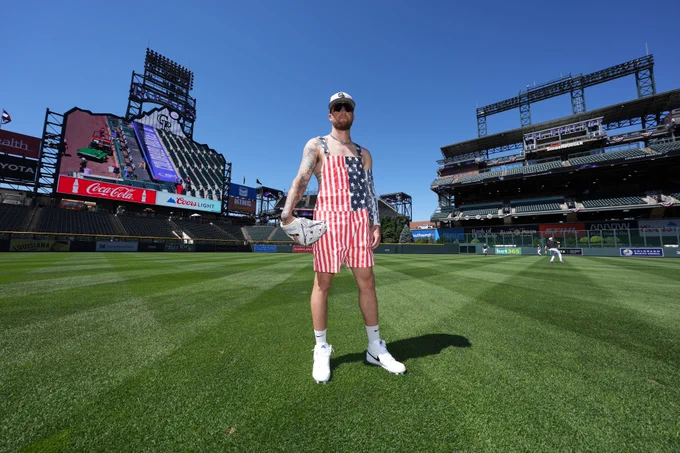 A photo of Kyle Freeland standing on the outfield grass at Coors Field. He is wearing short overalls with the American Flag pattern. He is wearing white cleats, and a white hat with the CR logo in American Flag print. 