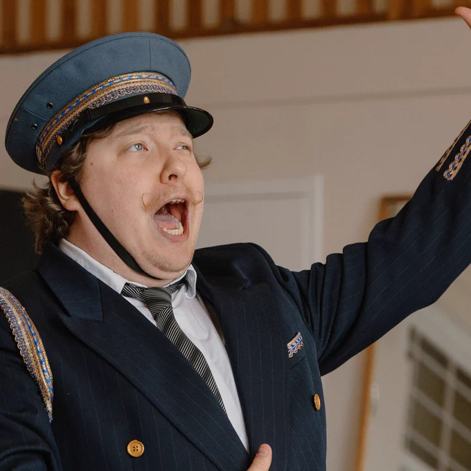A performer dressed in a navy blue suit and cap, passionately singing or acting with one arm raised and an expressive look on their face, in a rehearsal space.