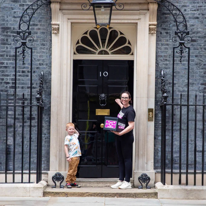Vic and Dylan stand outside the black front door of Number 10, Downing Street. They are knocking on the door and holding a box which contains a petition for Young Lives vs Cancer's campaign  Running On Empty. Vic is wearing a black Young Lives vs Cancer t-shirt and Dylan's white t-shirt has multi-coloured dinosaurs on it. They are both looking at the camera and smiling while they knock