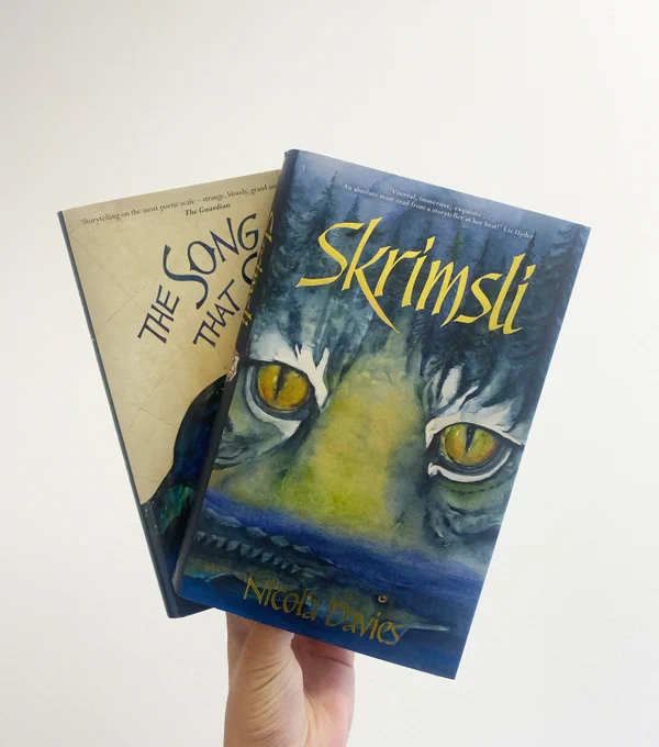 Hand holding two hardback books up to a white background. Book one is The Song That Sings Us and book two is Skrimsli. Both YA fantasy written by Nicola Davies and illustrated by Jackie Morris.