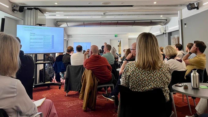 A conference room with a large group of people looking at a big screen with projected text on it.