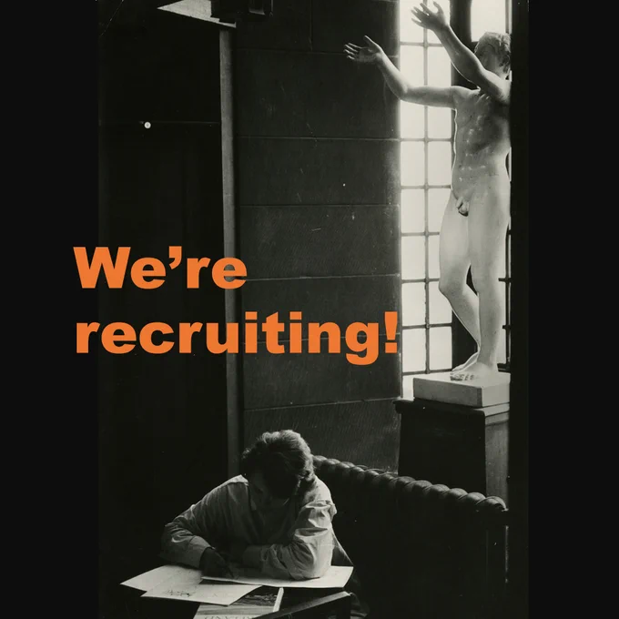 Black and white photograph of a person reading papers at a desk in the Mackintosh library, overlooked by a plaster cast nude. Orange text overlays the image, reads "we're recruiting!"