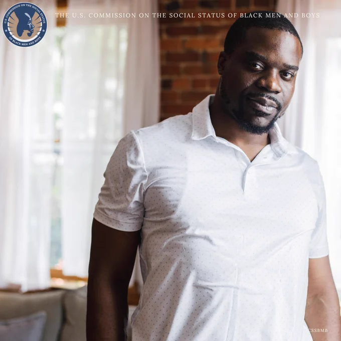 Black male in white buttoned shirt looks into camera while standing in living room. 