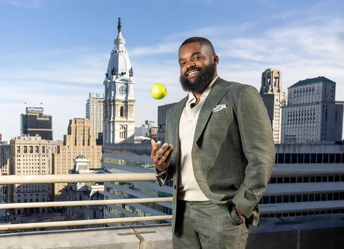 Man in a suit stands on a rooftop with cityscape background, smiling and tossing a green apple in the air.