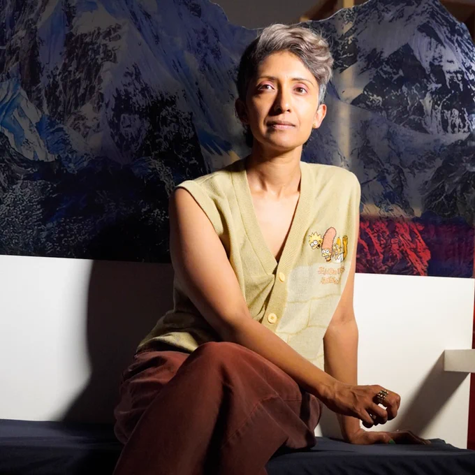 A brown-skinned woman with short grey hair, sat crossed-legged on a white bench, wearing a beige button down vest and rust coloured trousers, a cut out of snow covered mountains behind her.