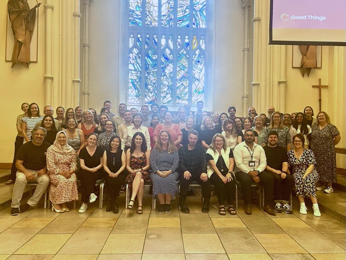 Staff and representatives from our board of trustees and ambassador National Digital Inclusion Network hubs. In the front row, people are sitting on chairs. In the rows behind, people are standing. There is a large stained glass window in the background.