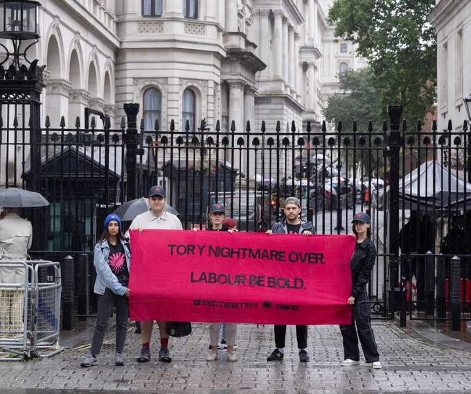 5 young people stand outside the gate to Downing Street, wearing hats and jackets to protect from the rain. They hold a pink banner that reads: Tory nightmare over, Labour be bold.