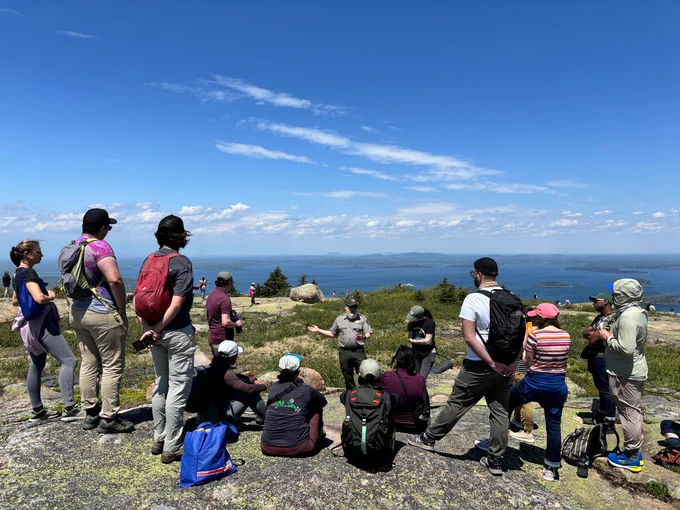 A group of Fellow's circled around a park ranger speaking atop a peak overlooking the ocean.