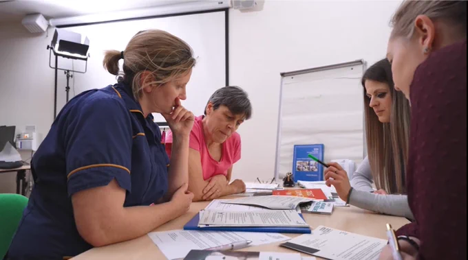 Nurse and three members of the public sit around a table looking at documents toghether on the table. 