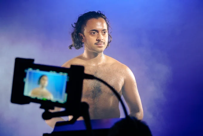 A brown-skinned person with a bare torso stands facing the camera, but looking to the side. They are in a studio with a purple background and a cloud of smoke behind them. A camera display screen is in the foreground but out of focus. 