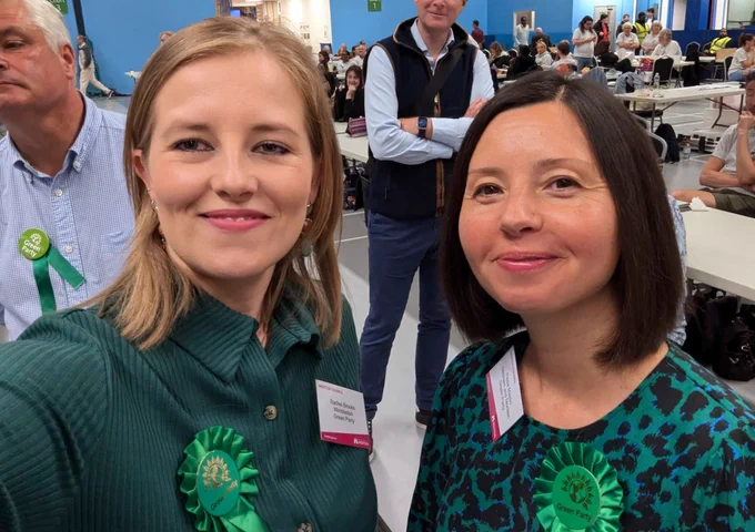 Selfie of our Wimbledon parliamentary candidate Rachel Brooks, and Mitcham and Morden parliamentary candidate and St Helier ward by-election candidate Pippa Maslin, at the count in Canons Leisure Centre, in the wee hours. Both are wearing green dresses and rosettes. Both are smiling. The counting tables, some of the counting staff, and other attendees of the event can be seen in the background.