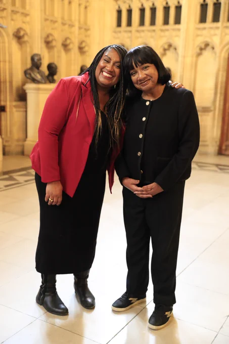 Bell Ribeiro-Addy MP and Diane Abbott MP stand together in the members' lobby in Parliament. Bell wears a red blazer and black skirt and Diane wears a black suit.