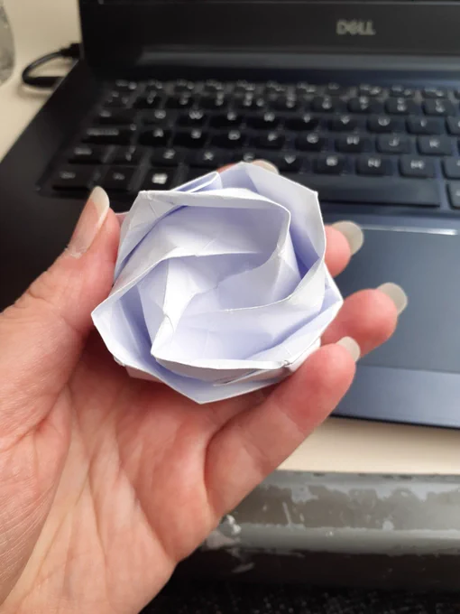 A white paper origami rose is held in a hand with a laptop in the background.