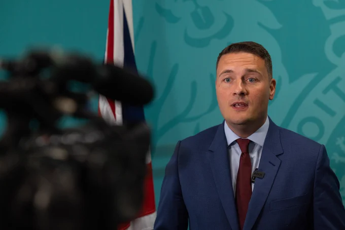 Health and Social Care Secretary Wes Streeting delivers a speech to camera standing in front of an official DHSC backdrop with the UK Government crest on it, alongside a flag of the UK.
