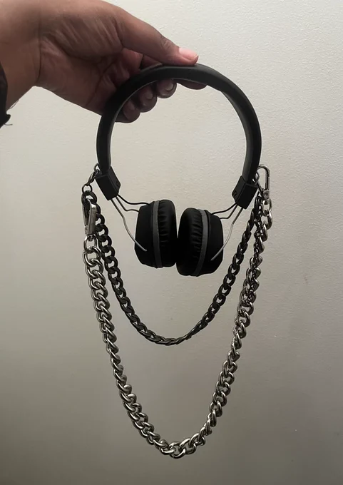 The hand of Bee, a Black person, holds her black & grey over the ear headphones, which have a silver chain & black rhodium chain attached, against a plain white background.