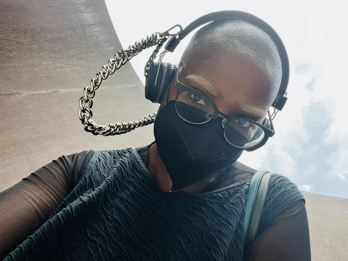 Selfie of Bee, a Black person with a shaved head & blonde eyebrows, wearing a black n95 respirator with glittery black eyeglasses, black & grey headphones with a silver chain & black rhodium chain attached, and a textured sleeveless black top over a mesh long sleeve black top. She’s standing inside of the sculpture Joe by Richard Serra and both the steel and sky behind her are slightly visible.