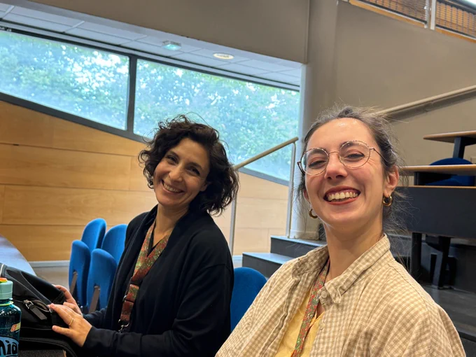 Dr Gershy sits smiling on the left side of the picture wearing a black cardigan and Eva sits n the right hand side with a very wide smile wearing glasses, a checked shirt and yellow dress.
