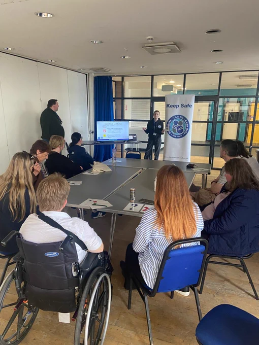 A group of people seated round a table receiving an input from a female who is stood in front of them, with a screen beside her that is displaying information