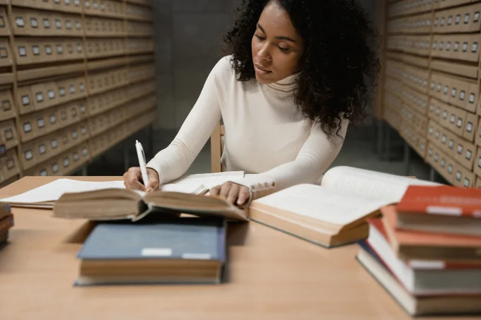 A person sits at a desk occupied by lots of books within a library archive, concentrating on writing notes as they look at an open book for information.