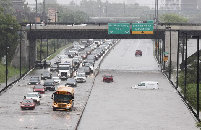 A flooded highway in Toronto with vehicles, including a school bus and a van, partially submerged in water. Cars are stuck in traffic due to the flooding under an overpass with green directional signs overhead.