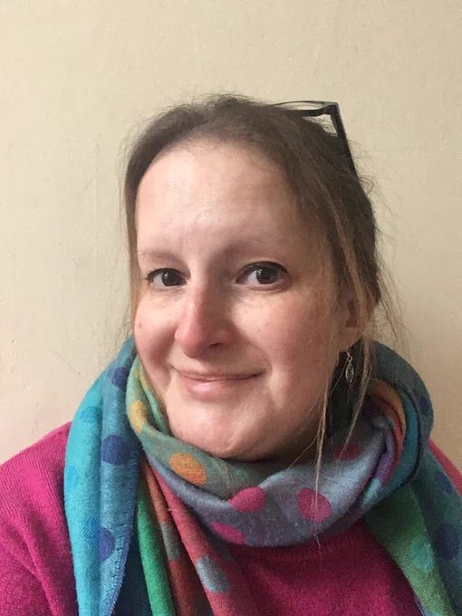 Steph is a white woman with long hair loosely pulled into a bun. She is smiling and wearing a colourful scarf and pink jumper. 
