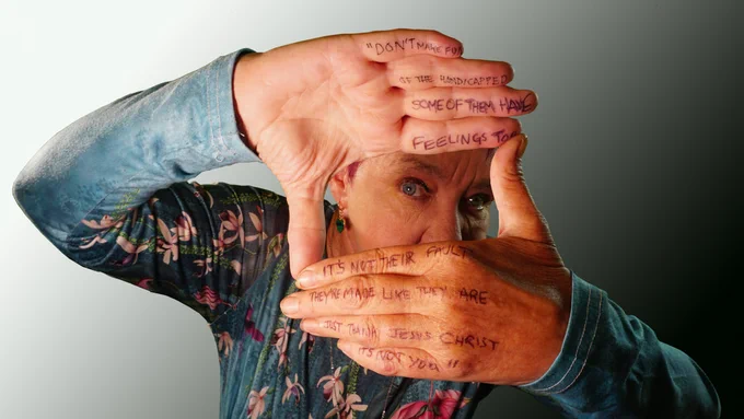 An image of Karen Sheader by Derek Matthews. Wearing a patterned denim shirt, Karen is holding her hands in a square as if framing an image and looking through. She has lyrics written on her hands including the phrase 'don't make fun of the handicapped some of them have feelings too' heavy with irony.  Also - 1It's not their fault, they're made like they are, just thank Jesus Christ it's not you.'