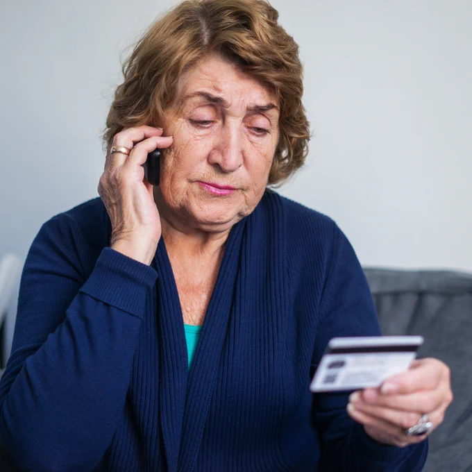 An older person sitting on a sofa, looking distressed while holding a credit card in one hand and a phone to their ear with the other hand.