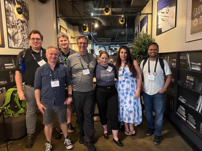 Seven people (5 men, 2 women) are standing together and smiling. 
Shiri, a light-skinned bronde is standing in the middle, and is wearing a T-shirt that bear the following writing:
DISABILITY
DISABILITY
**DISABILITY**
DISABILITY
DISABILITY
DISABILITY 
is not a bad word