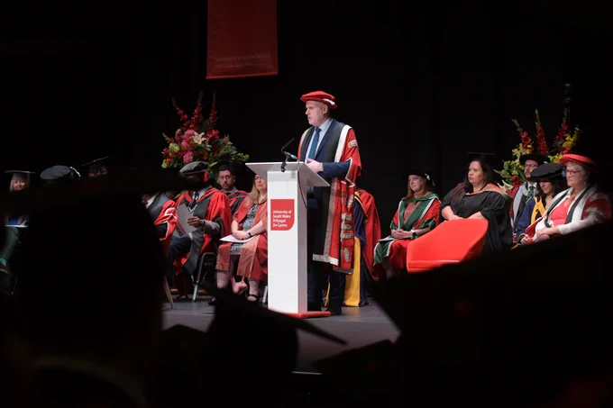 The image shows a graduation ceremony at the University of South Wales. A person in academic regalia is speaking at a podium labeled with the university's name. Other faculty members, also in academic robes, are seated on stage, with some wearing caps and gowns. The background features a floral arrangement and a banner with the university's name. The audience, partially visible, is in the foreground wearing graduation caps.


