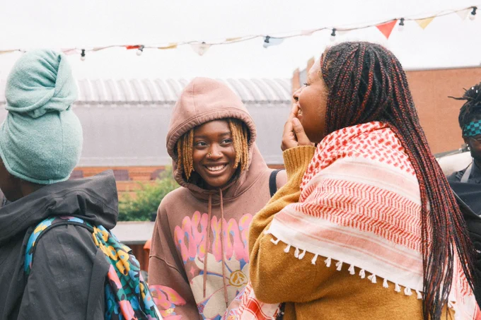 Solidarity Economy Showcase attendees laughing outside. One person has a hoodie on with their hood up, while the other person is wearing a red keffiyeh. Bunting is visible in the background.