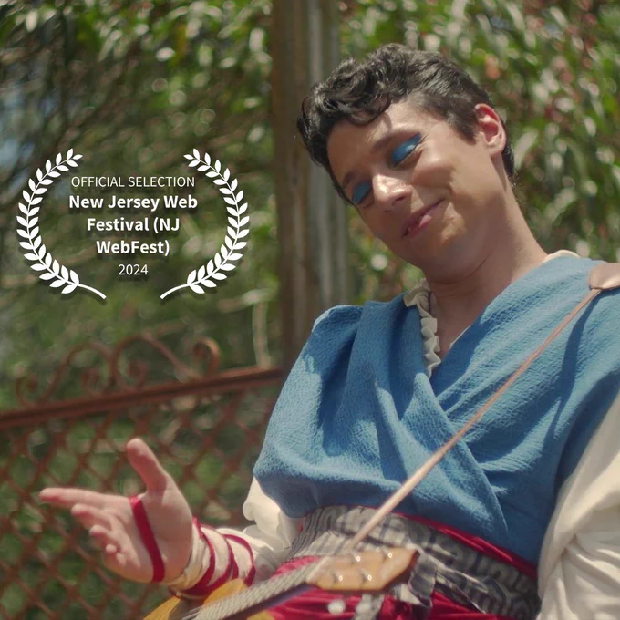 Vic sorrindo de olhos fechados satisfeito, sob o sol do jardim de Lavin. Ele faz um gesto com a mão que parece indicar os Louros em que se lê: official selection New Jersey Web Festival (NJ WebFest) 2024
