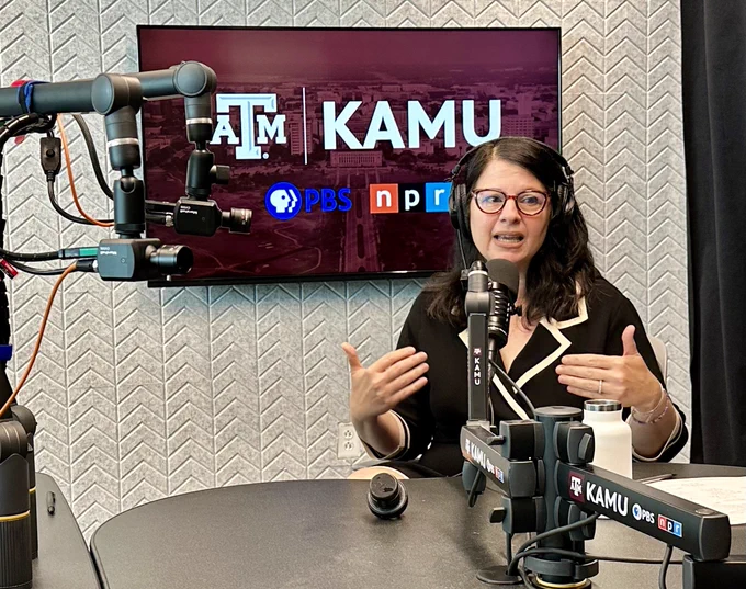 A person is seated at a broadcast studio desk, gesturing while speaking into a microphone. Behind the individual is a screen displaying logos including "Texas A&M", "KAMU", "PBS", and "npr". The studio is equipped with various broadcasting microphones and technological equipment.