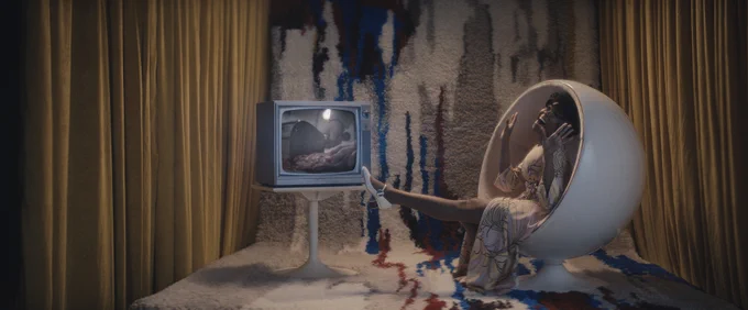 Moses Ingram stars as Cleo Johnson in Apple TV ’s Lady in the Lake. Moses is sitting in a ball-like white chair, wearing a light pink patterned dress and white heels. Her left foot is being kicked into the air and her hands are held up as she looks up at the ceiling with a big smile on her face. The chair she is sitting on is sitting on top of a red, blue, black, grey and white patterned carpet, that extends all the way up into the wall behind her. On the left and right side of her are mustard yellow curtains. Placed next to the chair is a small white table with an old-school TV from the 60s sitting on top of it. On the screen there is an image of a piece of raw meet with a butcher knife going through it. 