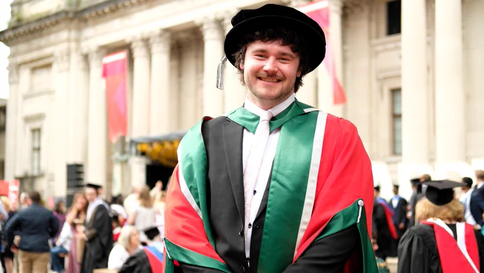 PhD graduate Joshua Moore standing in front of Glamorgan Building wearing his red and green robes and black velvet bonnet.