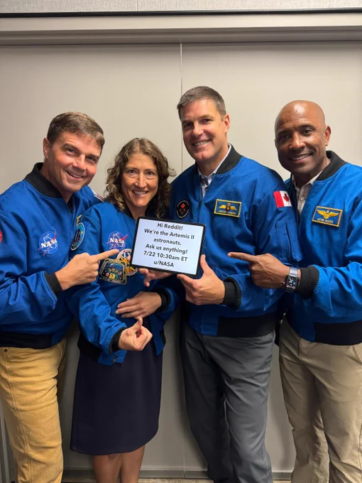The four astronauts of Artemis II — Reid Wiseman, Christina Koch, the Canadian Space Agency's Jeremy Hansen, and Victor Glover — stand and face the camera while holding a sign promoting NASA's Artemis II Reddit AMA, to be held on July 22 in the r/IAmA subreddit. The astronauts are wearing matching blue jackets and pointing at the sign. Credit: NASA