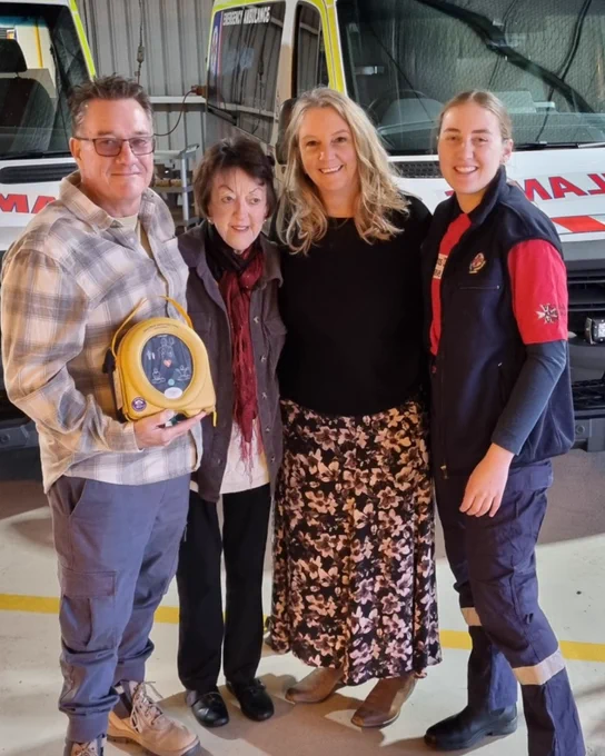 Four people standing in front of two emergency vehicles in a garage, smiling at the camera. One person is holding a defibrillator unit.