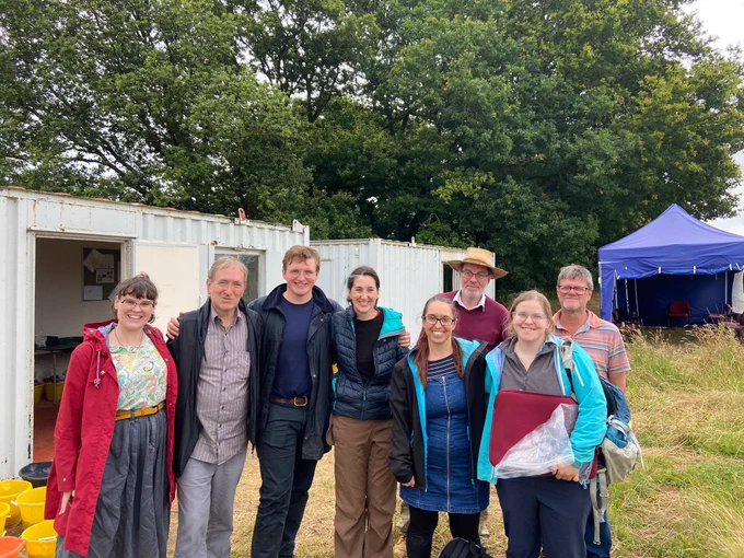 Eight members of the Anthropology team at the Natural History Museum stand in front of a white shed in an open field. Yellow buckets filled with dirt and mud can be seen in the lower left corner. There is a blue tent and many large oak trees in the background.