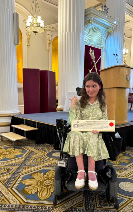 Anna, a young white woman in an electric wheelchair, posed pointing and side-eyeing a low wooden stage in an elaborately decorated  room.