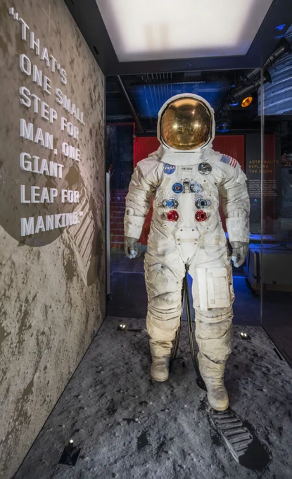 Neil Armstrong's Apollo spacesuit, positioned upright on a moon-like surface within an exhibit. Next to the suit, a wall features the quote: "That's one small step for man, one giant leap for mankind.